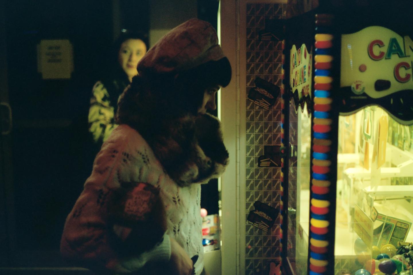 A woman in a hat and fur collar looks into a brightly lit claw machine while another woman stands in the background.