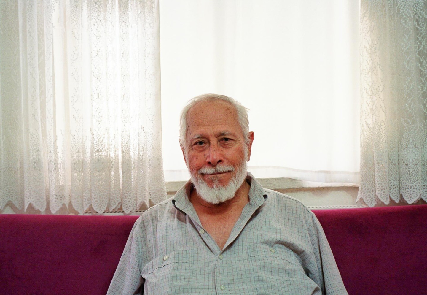 A man with a white beard sits on a purple couch in front of a window with white lace curtains.