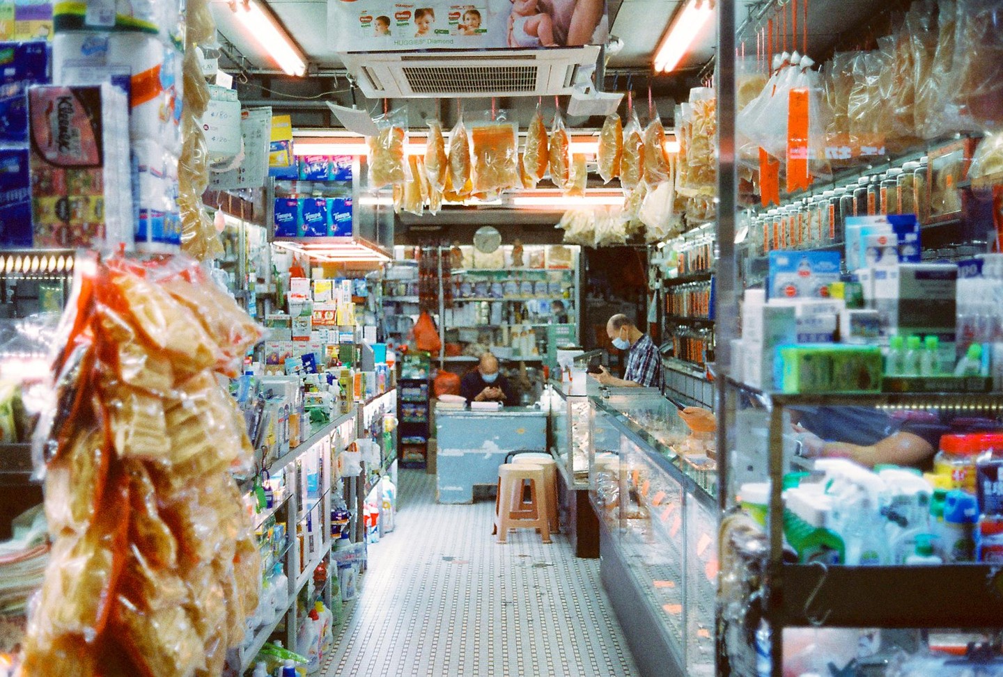 Two men wearing face masks are visible inside a narrow, brightly lit pharmacy filled with shelves of products and hanging packaged goods.