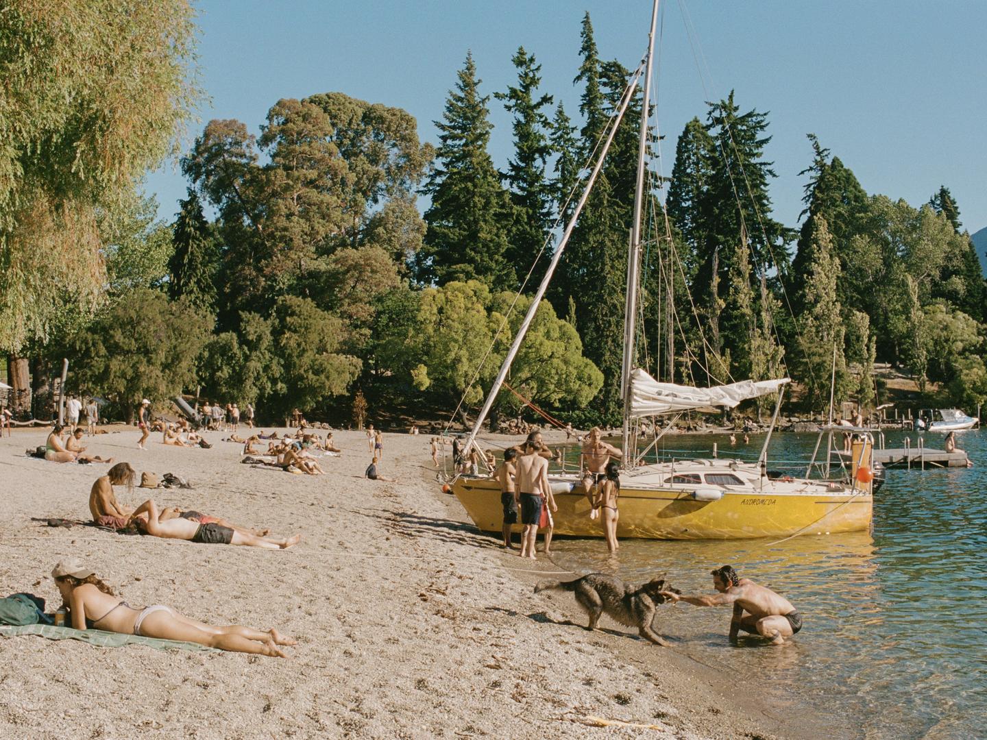 A man plays with a dog in the water next to a yellow sailboat on a crowded beach.
