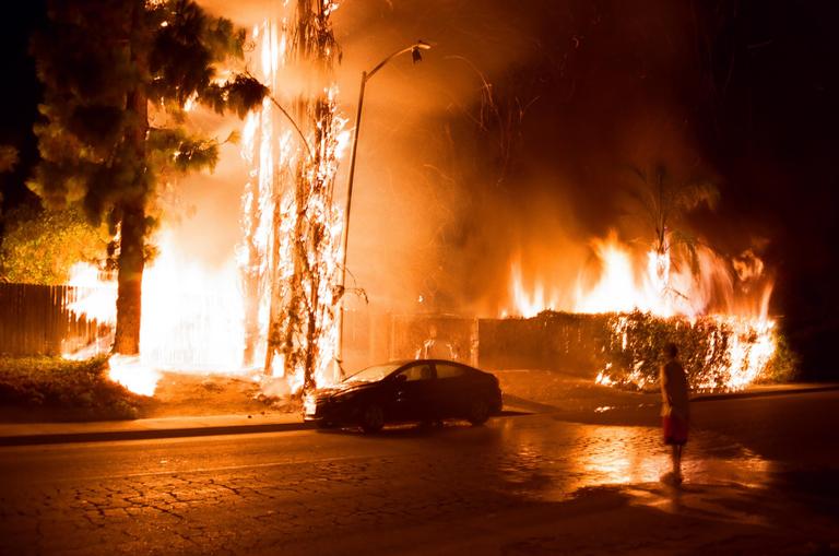 A person stands on a street at night, observing a car parked near a building and trees engulfed in flames.