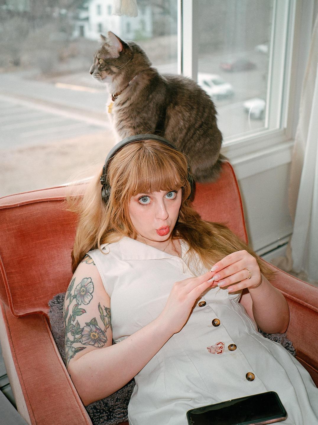 A woman with headphones sits in a chair making a funny face while a cat looks out the window behind her.