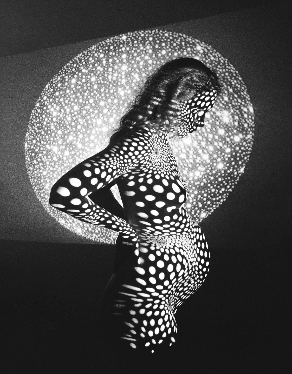 A pregnant woman is covered in projected polka dots, with a large polka dot circle projected behind her.