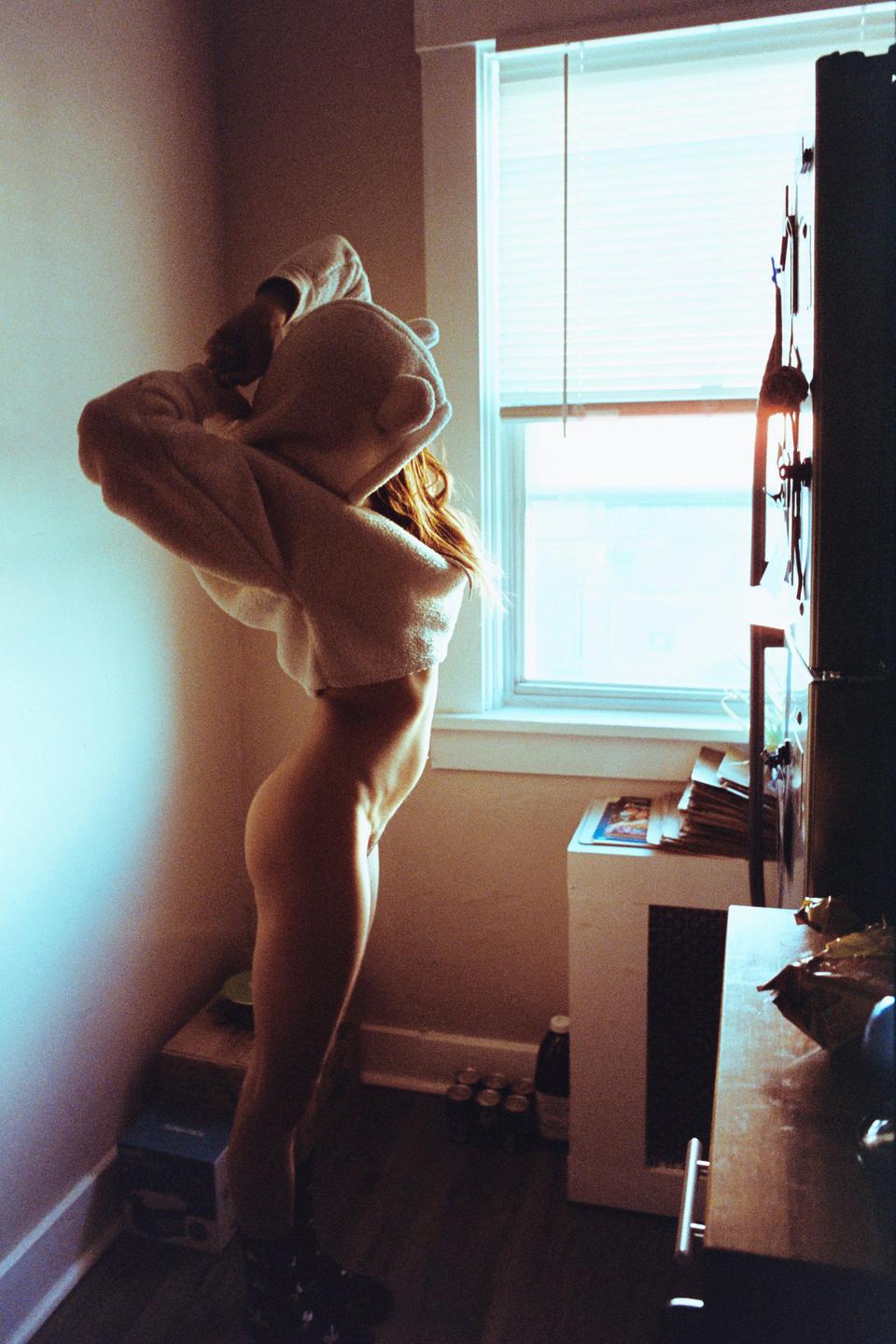 A woman stands partially dressed in a room with a window, illuminated by natural light.