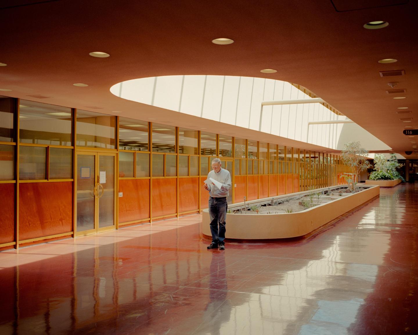 A man stands in a long hallway with a planter and reads a document.