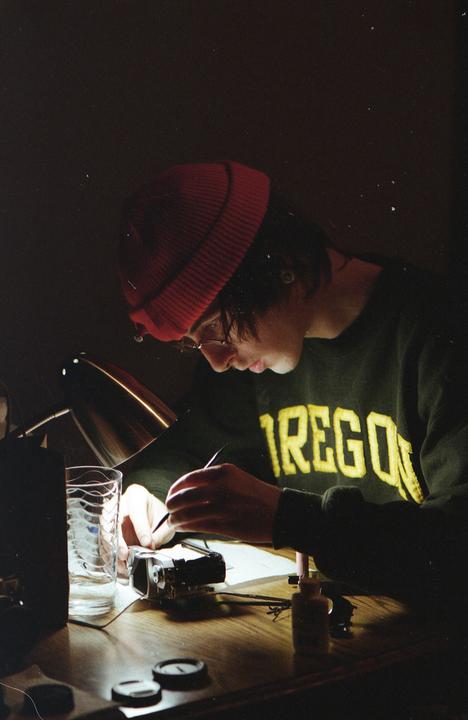 A person wearing a red beanie and a green sweatshirt with "OREGON" written on it is working on a camera at a wooden desk, illuminated by a desk lamp.
