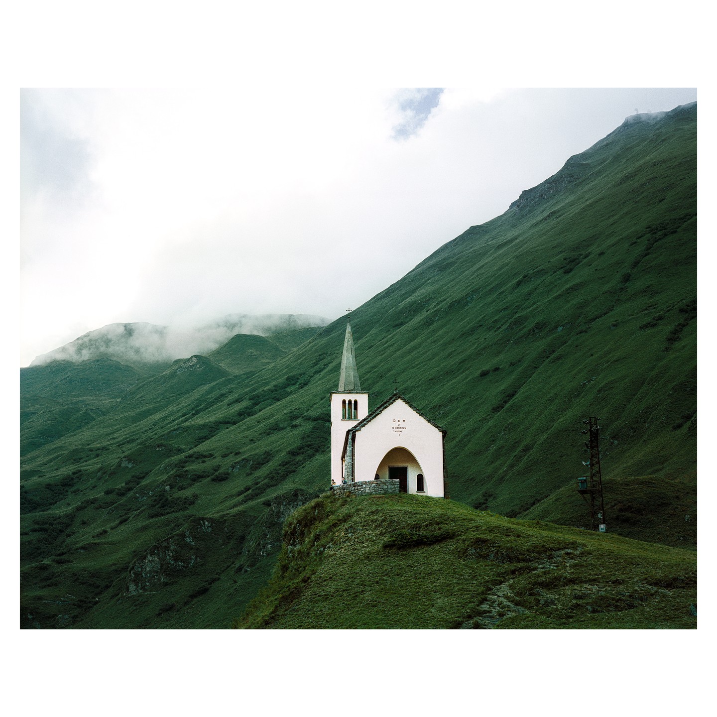 A small white church with a tall steeple stands on a grassy hill overlooking a valley with mountains in the background.
