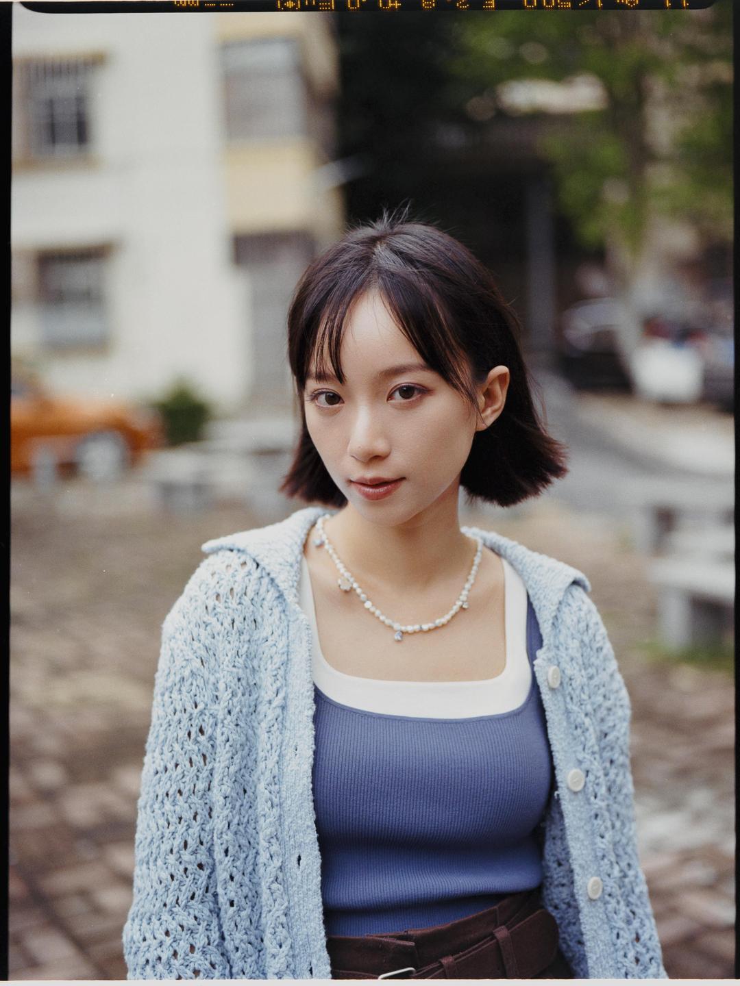 A woman with short dark hair and a pearl necklace looks directly at the viewer.