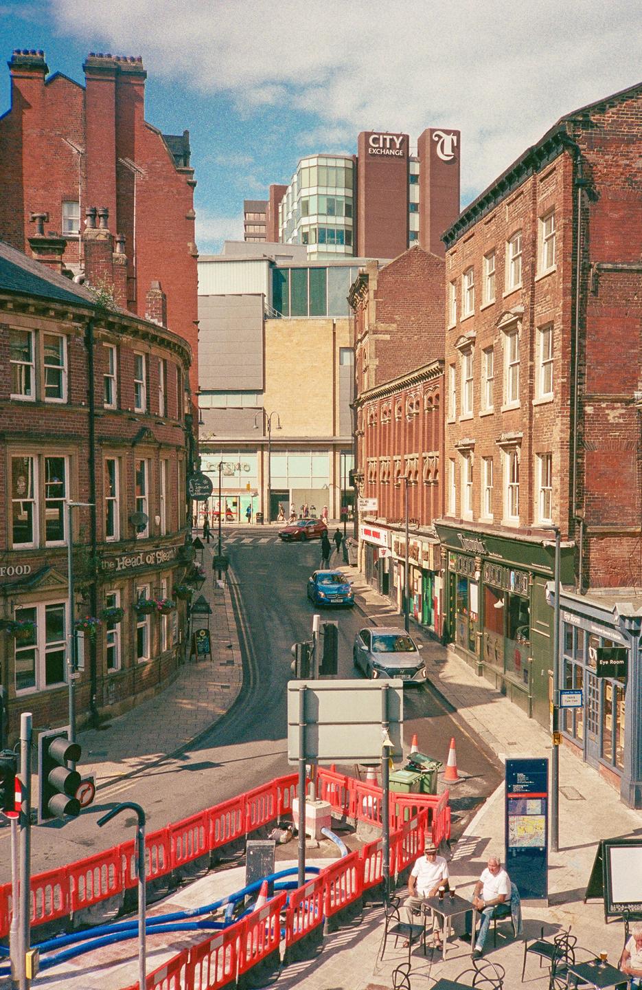 A street in Leeds, England, is lined with brick buildings and cars, with two men sitting at a table in the foreground.