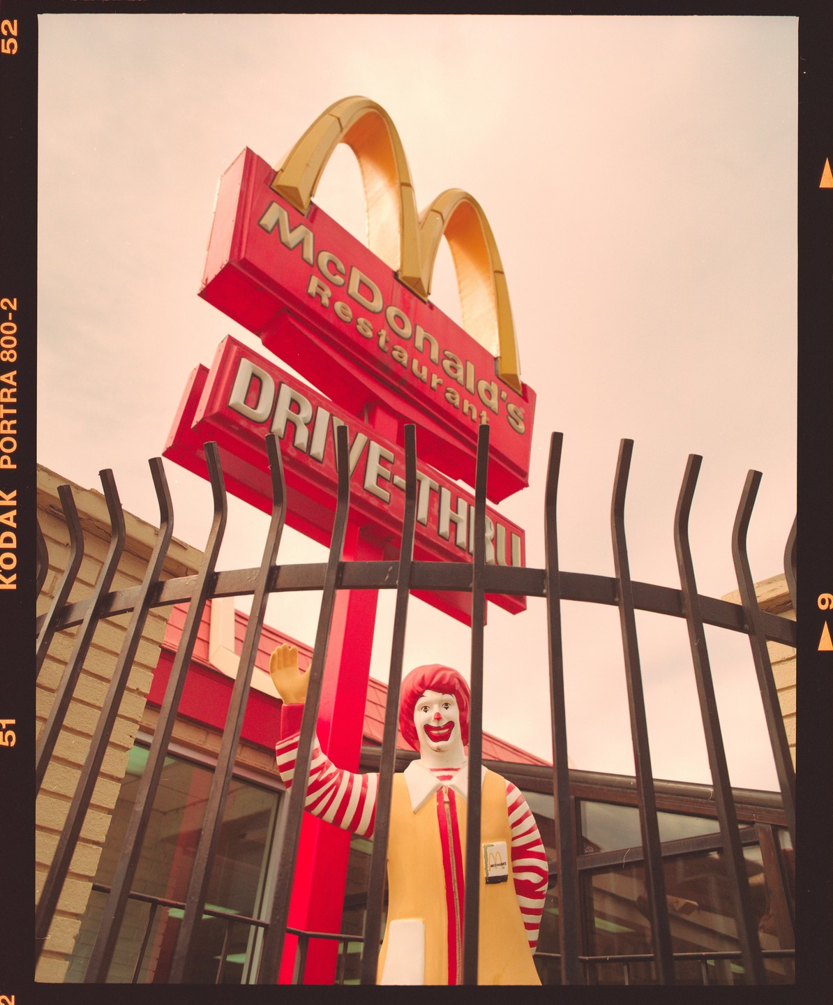 A Ronald McDonald statue stands behind a black metal fence with a McDonald's sign and golden arches in the background.