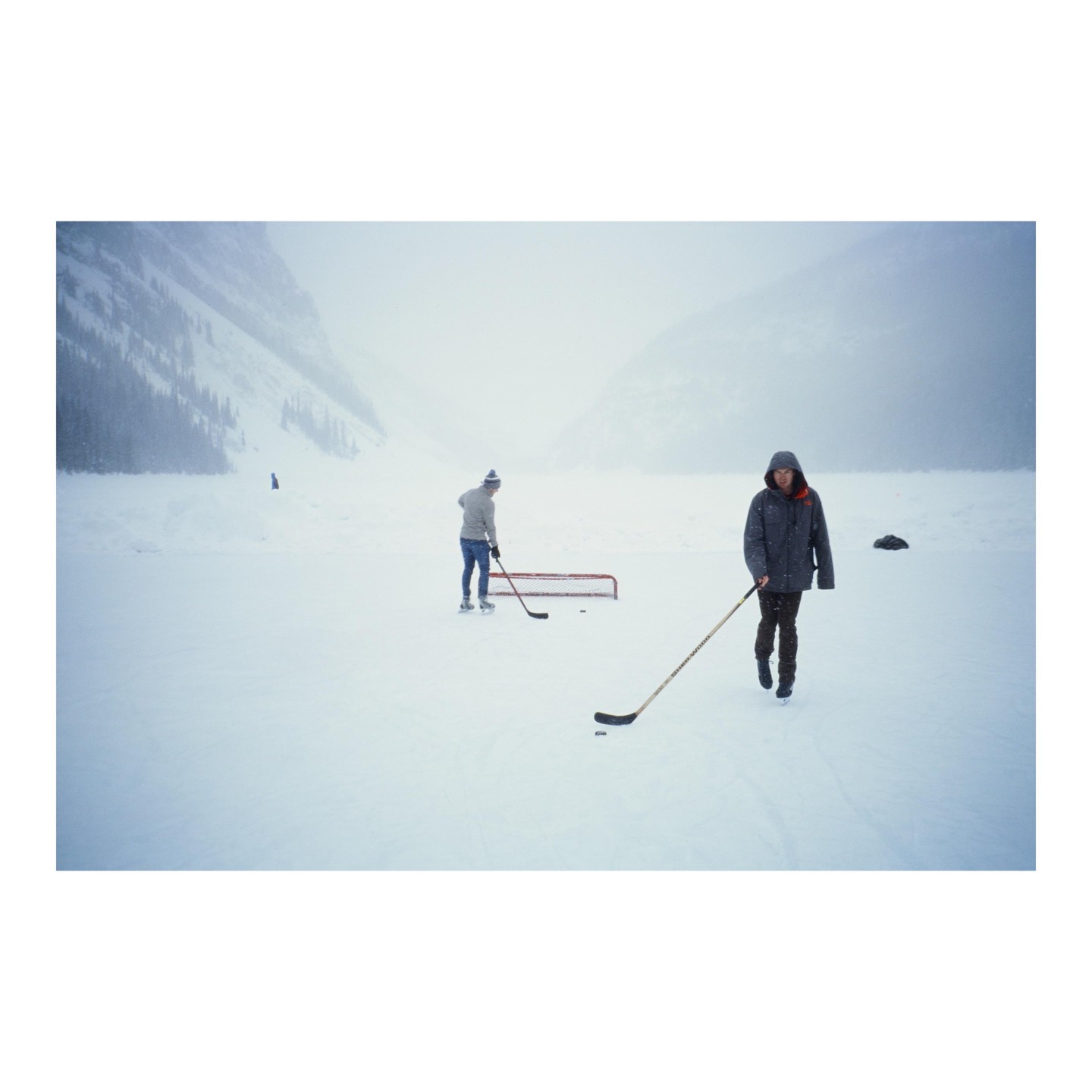Two men play hockey on a frozen lake surrounded by mountains in a snowstorm.