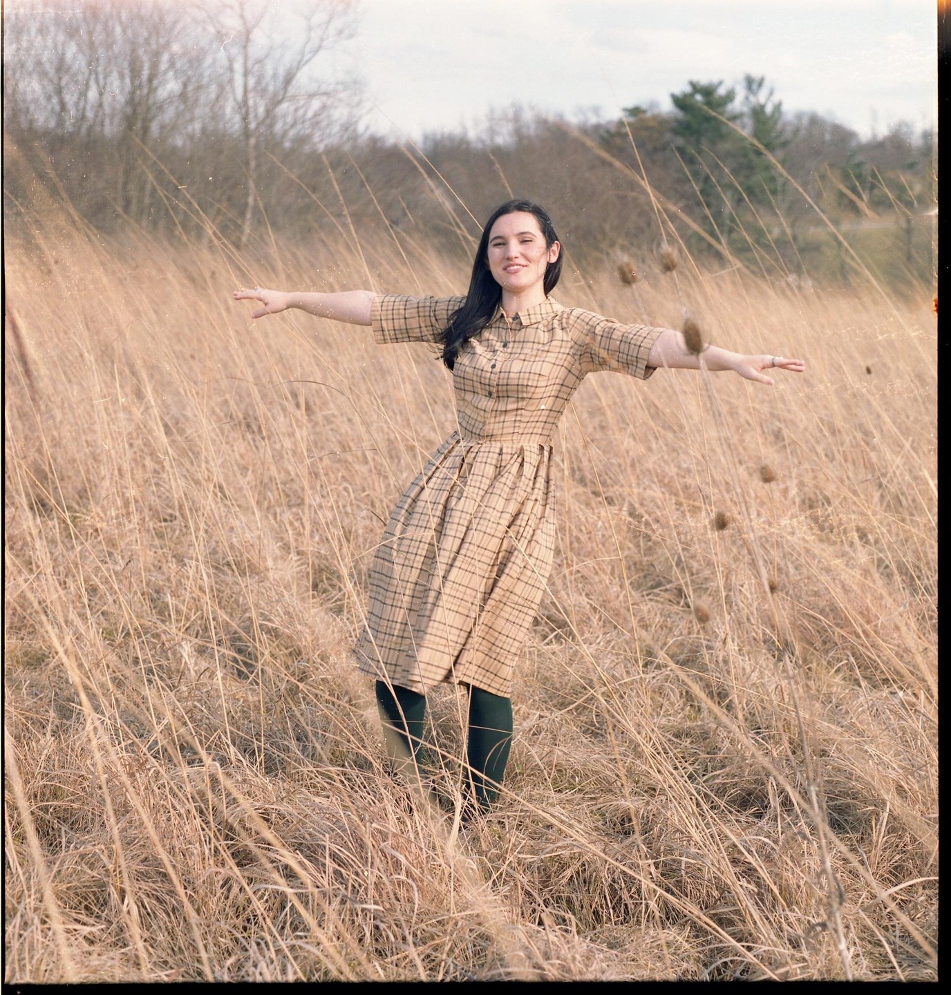 A woman in a plaid dress stands with her arms outstretched in a field of tall, dry grass.