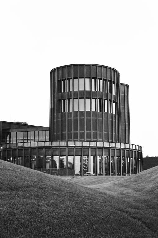 A modern cylindrical building with many windows is surrounded by grassy hills.