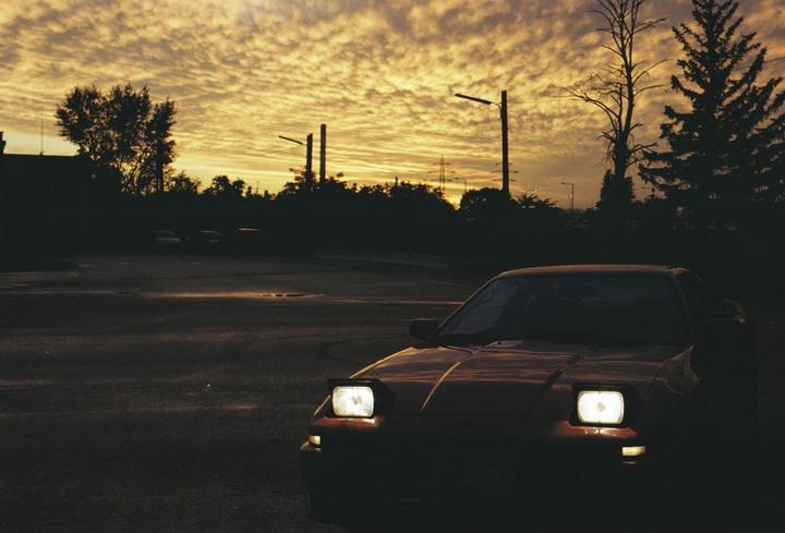 A car with its headlights on is parked in a lot at sunset.