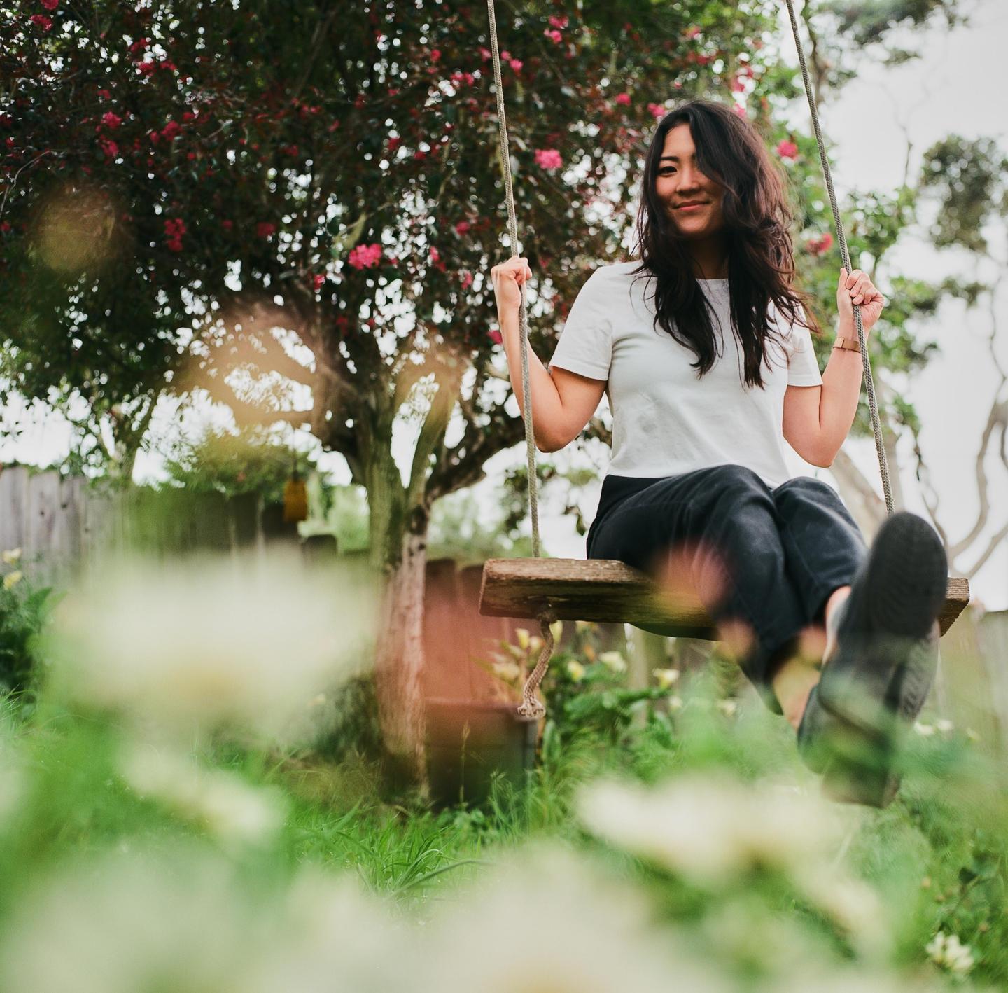 A woman sits on a wooden swing in a garden with a flowering tree in the background.