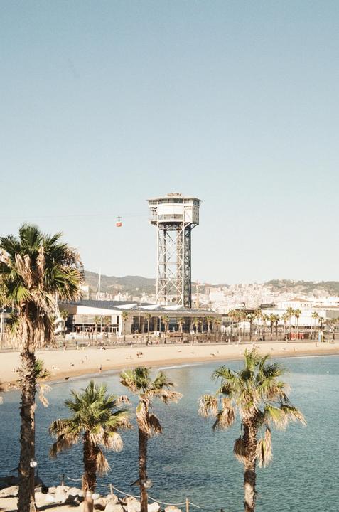 A cable car tower stands tall over a beach with palm trees and a city in the background.