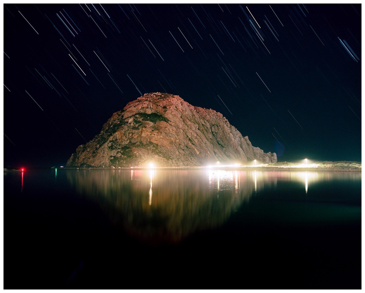 A large rock formation is illuminated at night with star trails visible in the sky above and lights reflecting on the water below.