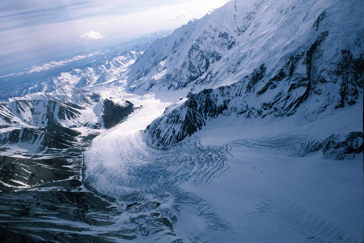 An aerial view shows a vast glacier winding through snow-covered mountains under a bright sky.
