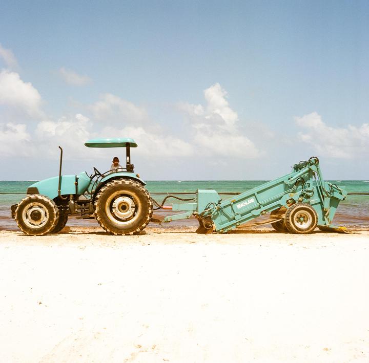 A teal tractor with a beach cleaner attachment is parked on a sandy beach next to the ocean under a blue sky with clouds.