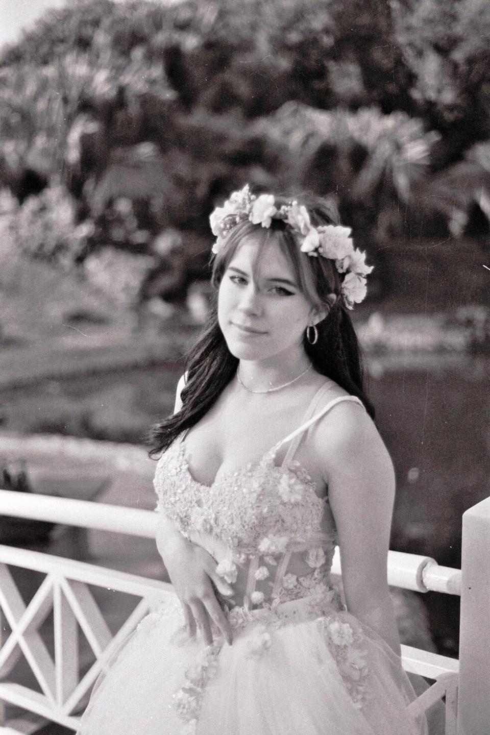 A young woman wearing a floral crown and an elaborate dress stands next to a white railing.