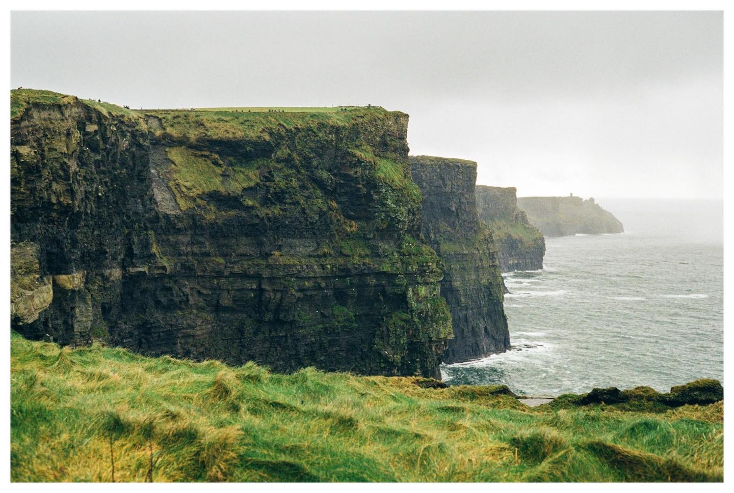 A dramatic landscape shows the Cliffs of Moher with green grass in the foreground and a hazy ocean.