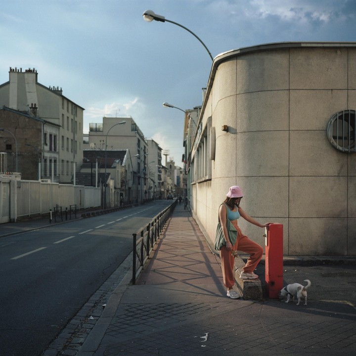 A woman in a pink hat and orange pants stands on a sidewalk next to a small dog and a red post.