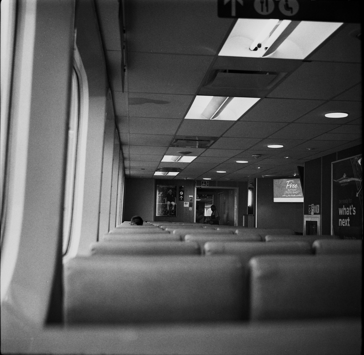 A long row of empty seats stretches down the aisle of a ferry with a few passengers visible in the distance.