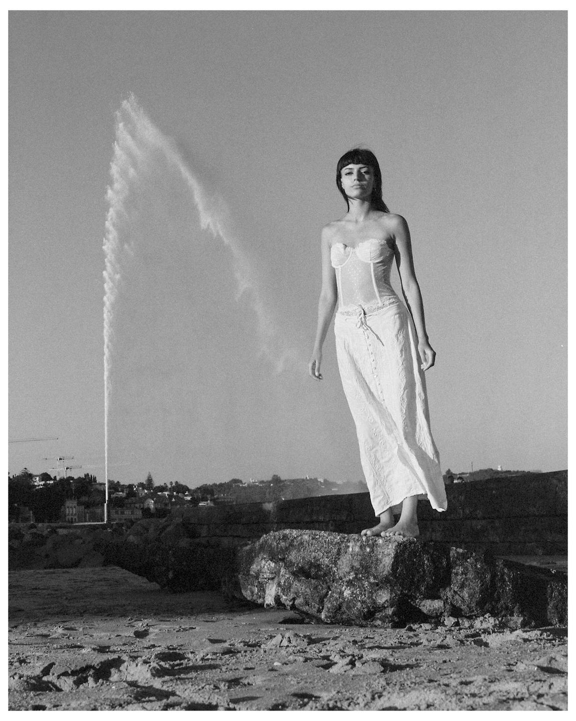 A woman stands barefoot on a rock on a beach with a fountain in the background.