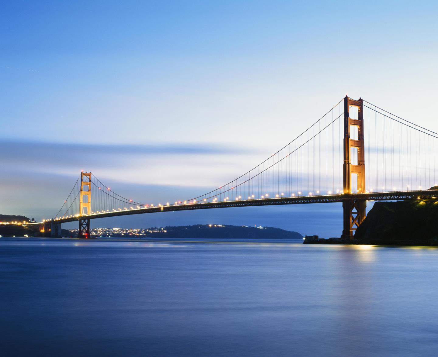 The Golden Gate Bridge is illuminated at dusk with city lights visible in the distance.