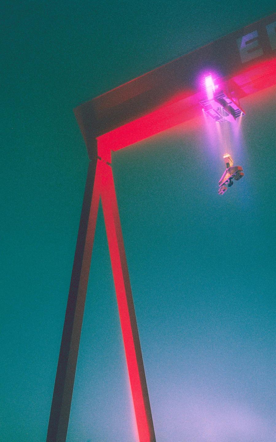 A large crane with red and purple lights stands against a teal sky.