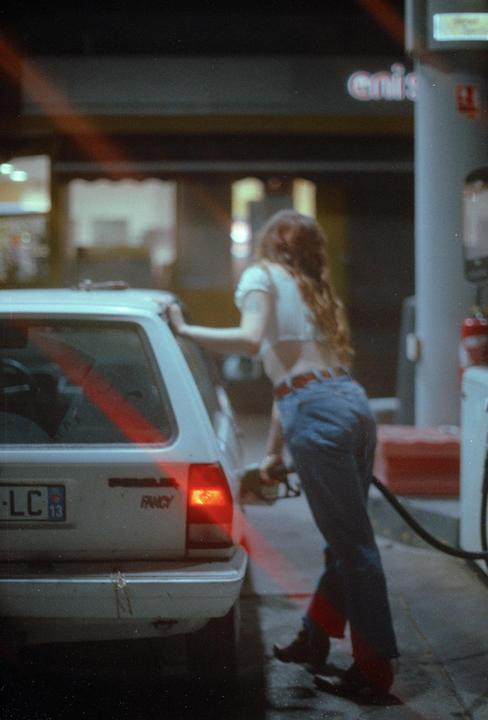 A woman with long hair is pumping gas into a white car at a gas station at night.
