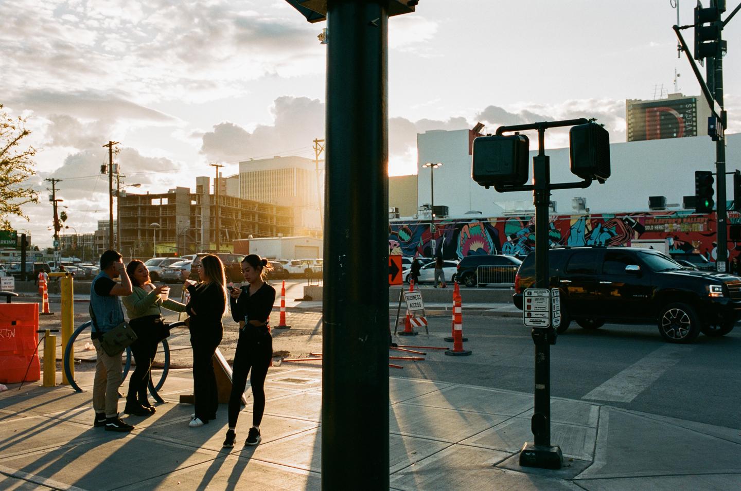 Four people stand on a sunlit street corner in Las Vegas with buildings and cars in the background.