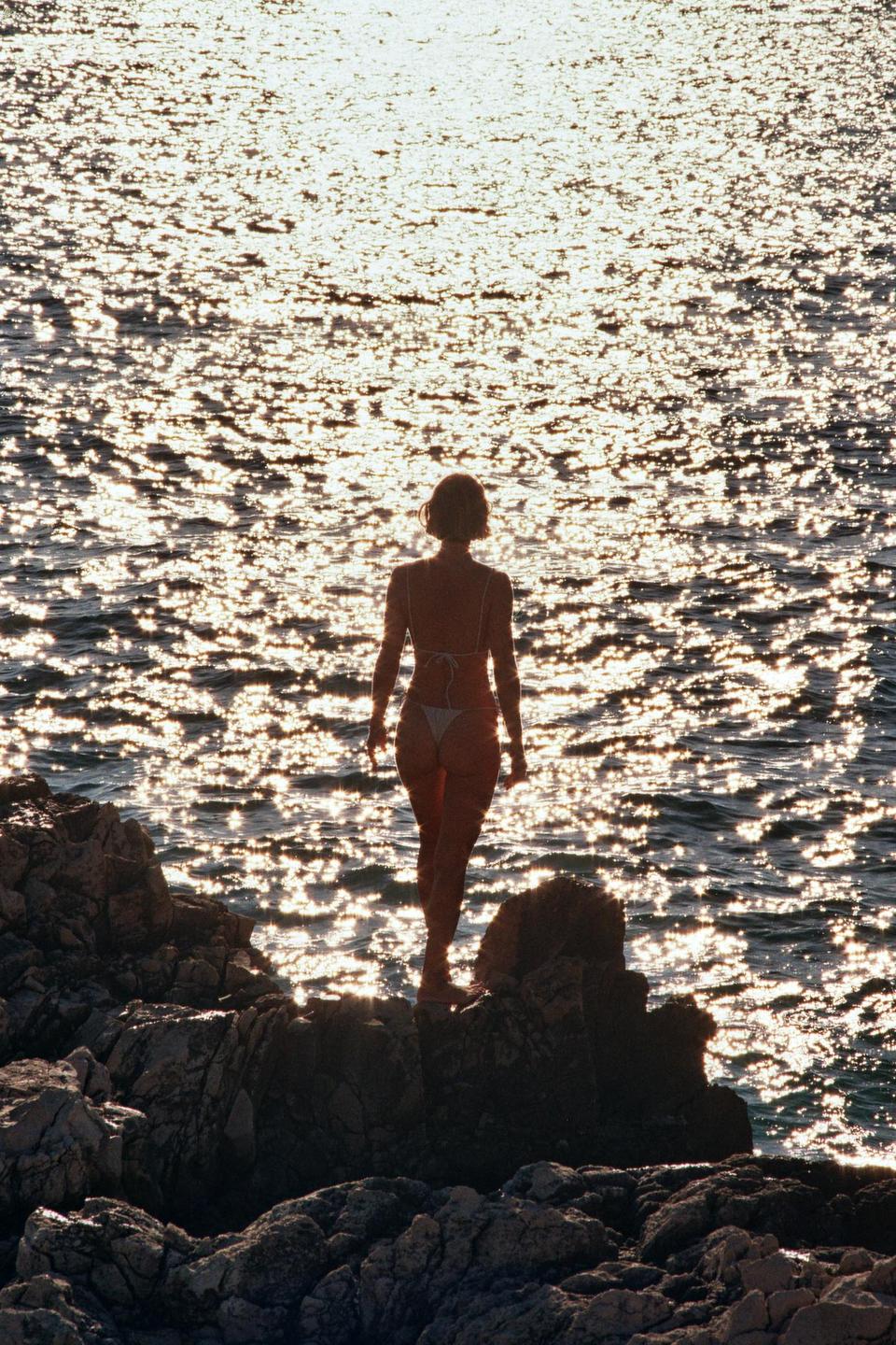 A woman in a bikini stands on rocks looking out at the sparkling water.