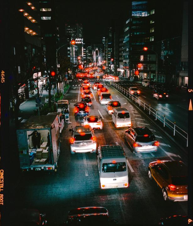 A busy street in Tokyo is filled with cars and trucks at night, with buildings lit up in the background.