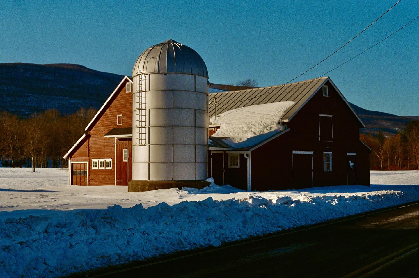 A red barn and a silo stand in a snowy field next to a road with mountains and trees in the background.