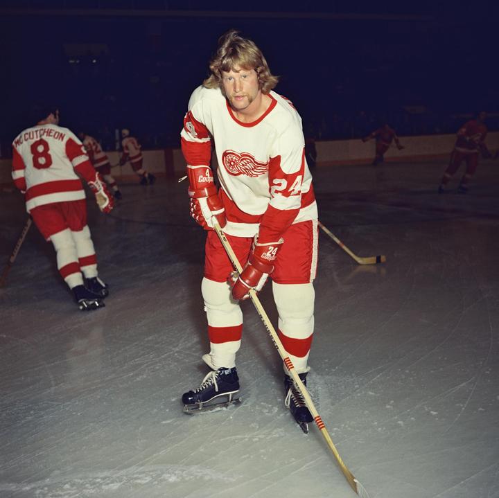 A man in a red and white hockey uniform stands on an ice rink holding a hockey stick.