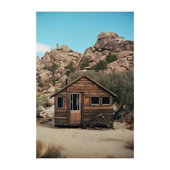 A small wooden cabin stands in a desert landscape with large rock formations in the background.
