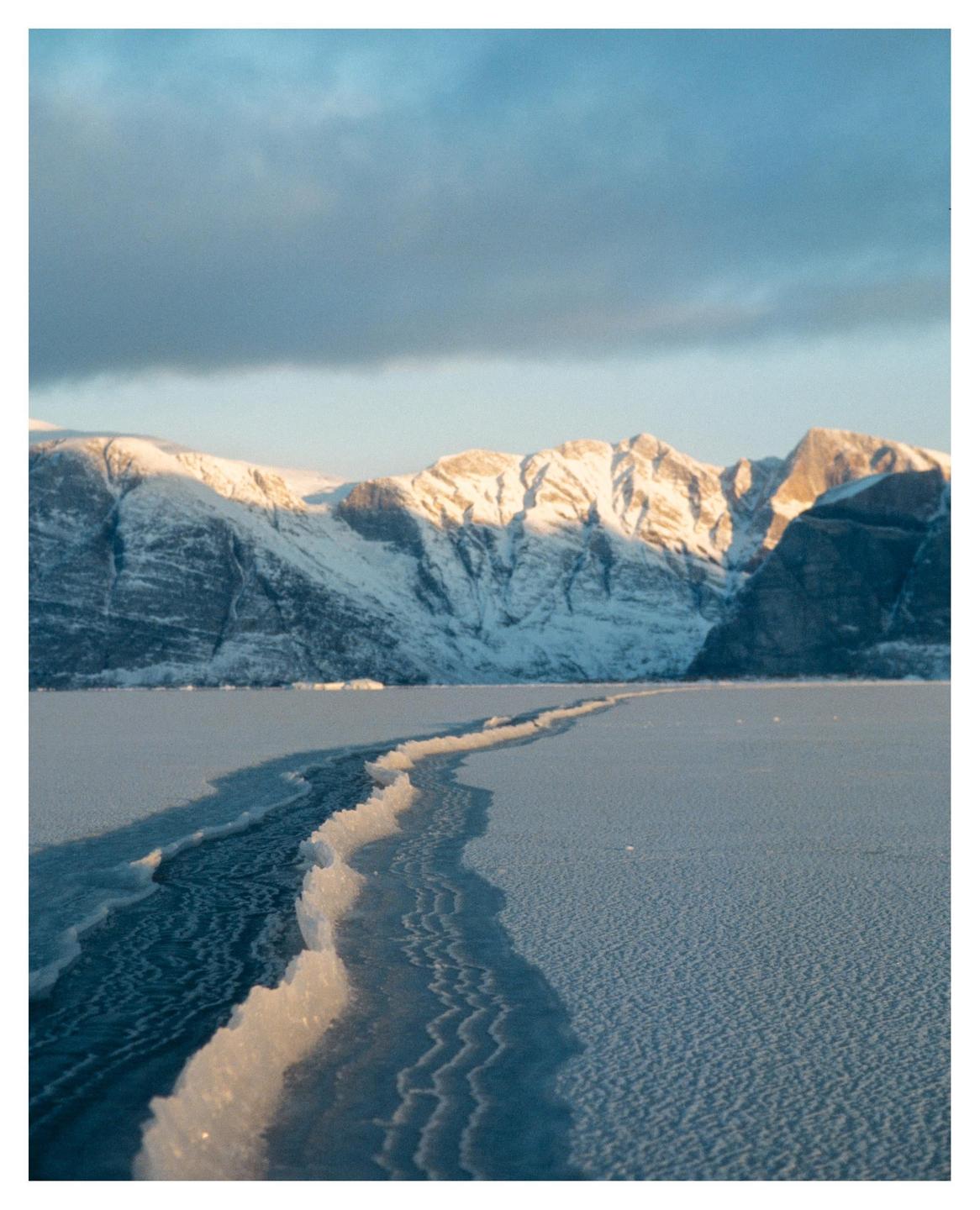 A crack in a frozen lake leads to snow-covered mountains under a cloudy sky.