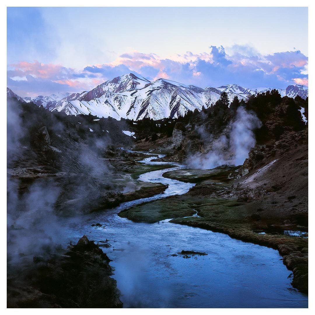A winding river flows through a valley with steam rising from the ground and snow-capped mountains in the background under a colorful sky.