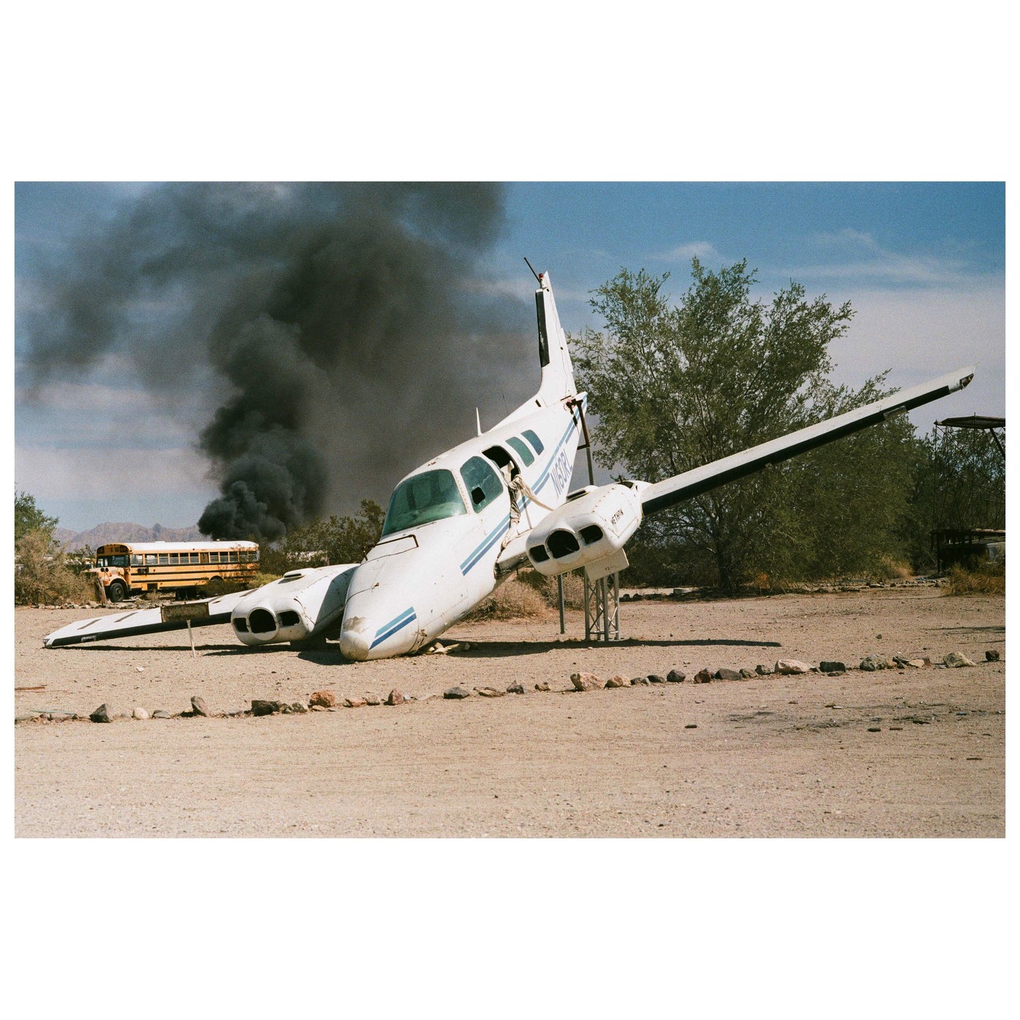 A white airplane wreck is on the ground with a school bus and black smoke in the background.