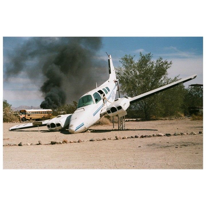 A white airplane wreck is on the ground with a school bus and black smoke in the background.