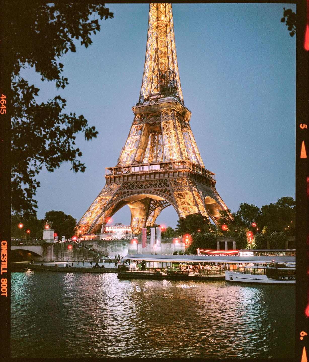 The Eiffel Tower is illuminated at night, towering over a river with boats and people on its banks.