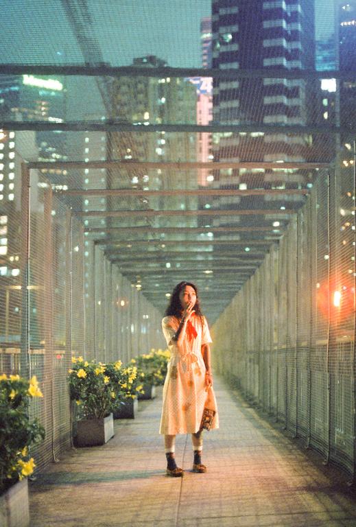 A woman stands in a covered walkway at night, surrounded by city lights and potted plants.
