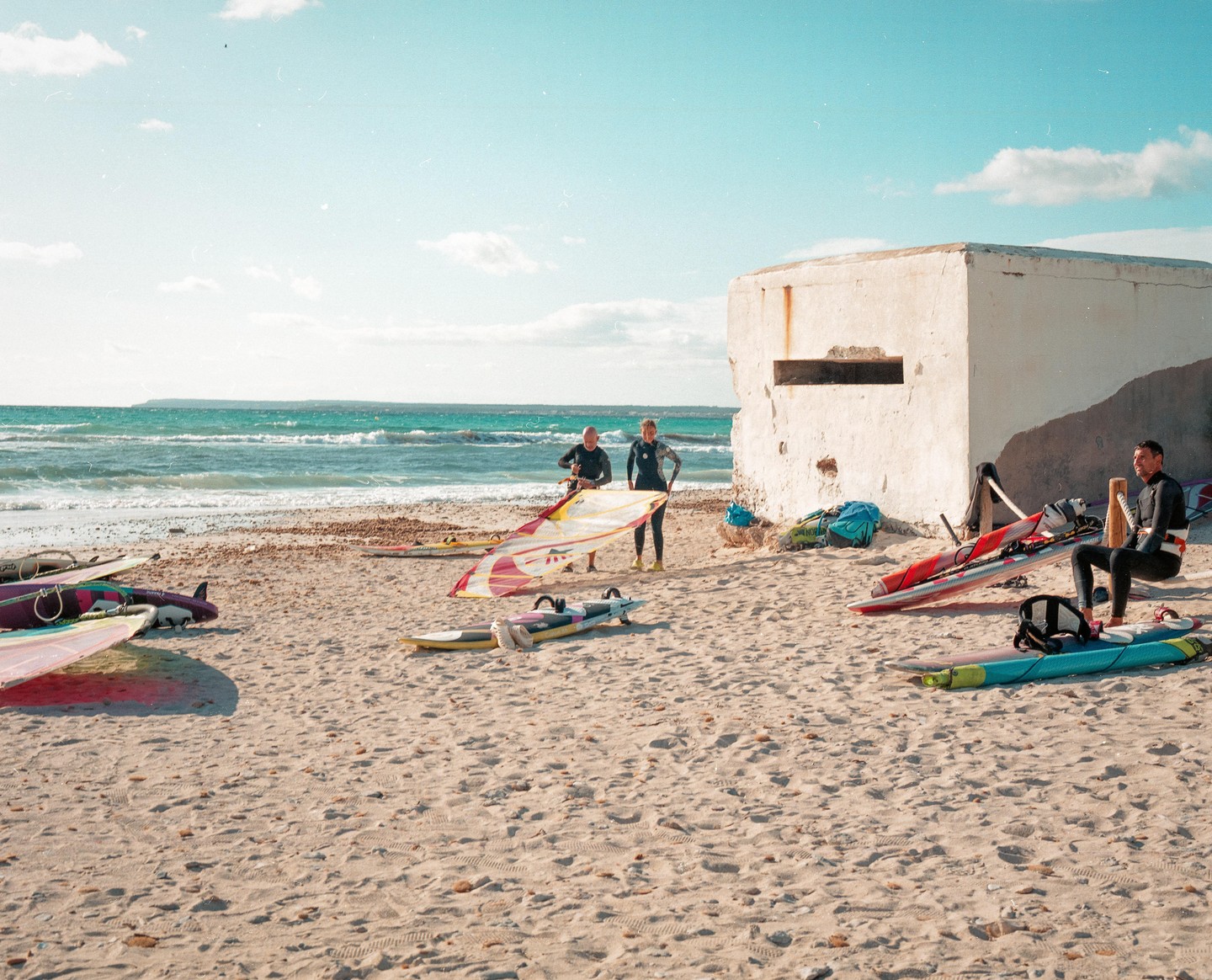 Three men in wetsuits are on a sandy beach with windsurfing equipment next to a concrete building.