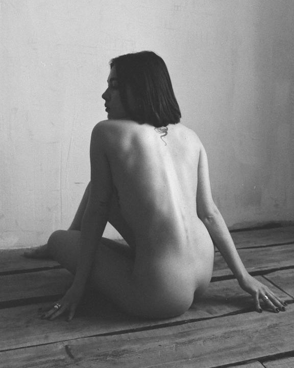 A woman sits on a wooden floor with her back to the viewer, looking over her shoulder.