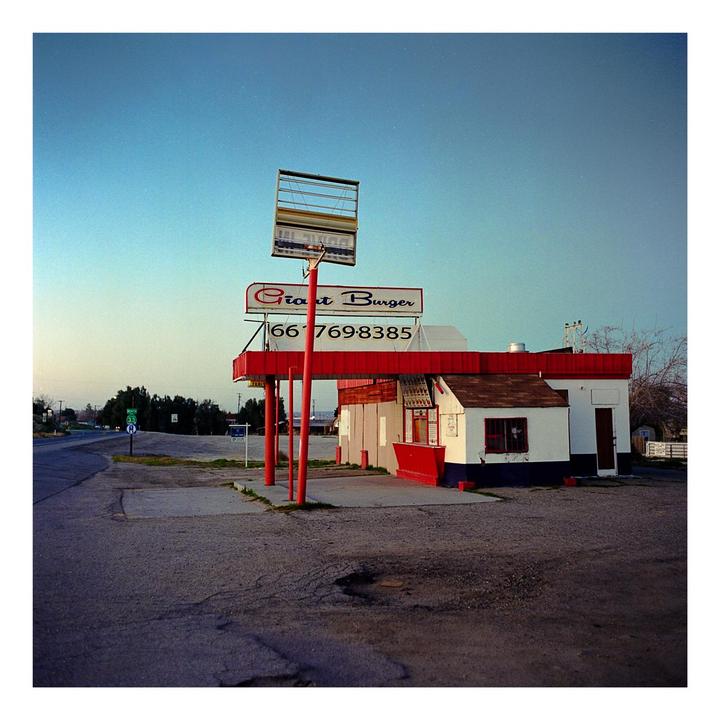 An abandoned Giant Burger stand with a large sign stands beside a road under a clear sky.