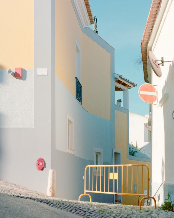 A yellow barrier stands on a cobblestone street between buildings with yellow and blue walls.