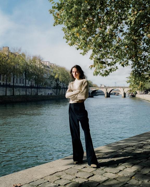 A woman stands on a cobblestone riverbank with a bridge and buildings in the background.