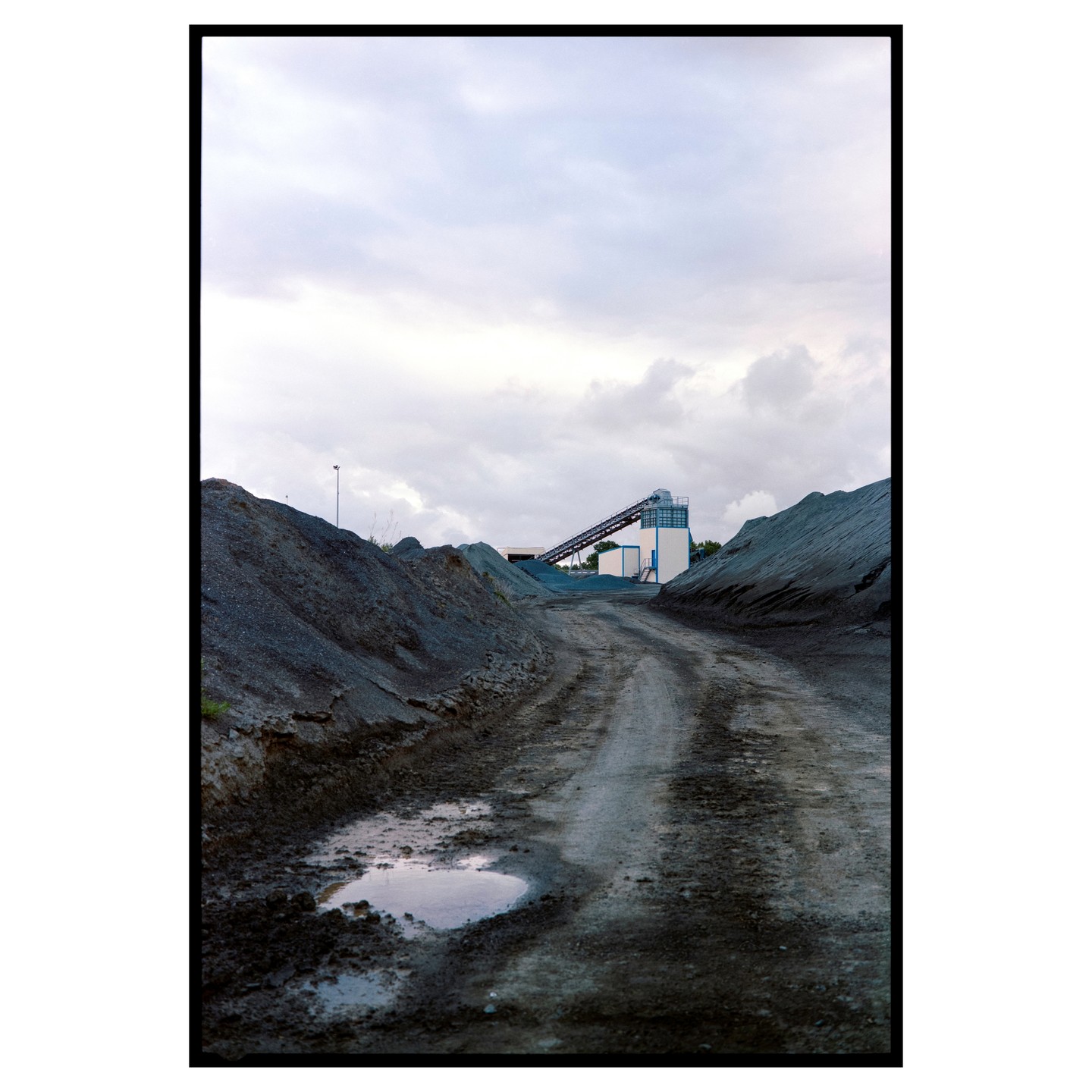 A dirt road with a puddle leads towards a quarry building and conveyor belt under a cloudy sky.