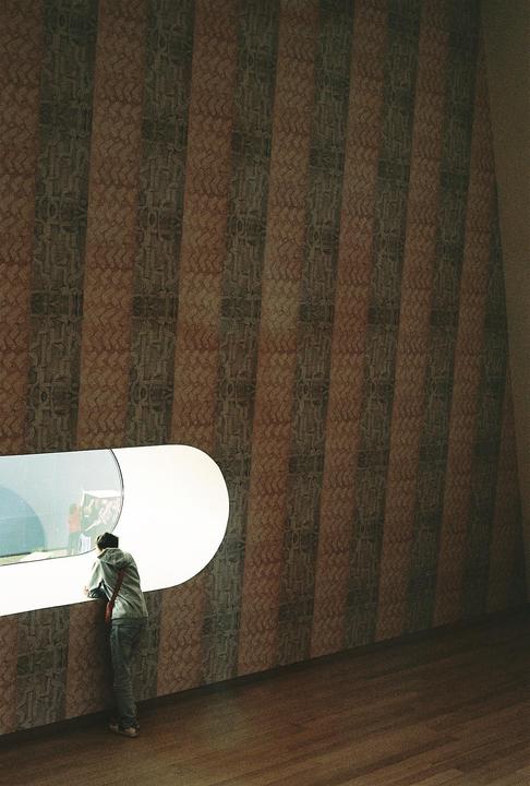 A person in a hoodie looks out a curved window in a room with patterned walls and a wooden floor.